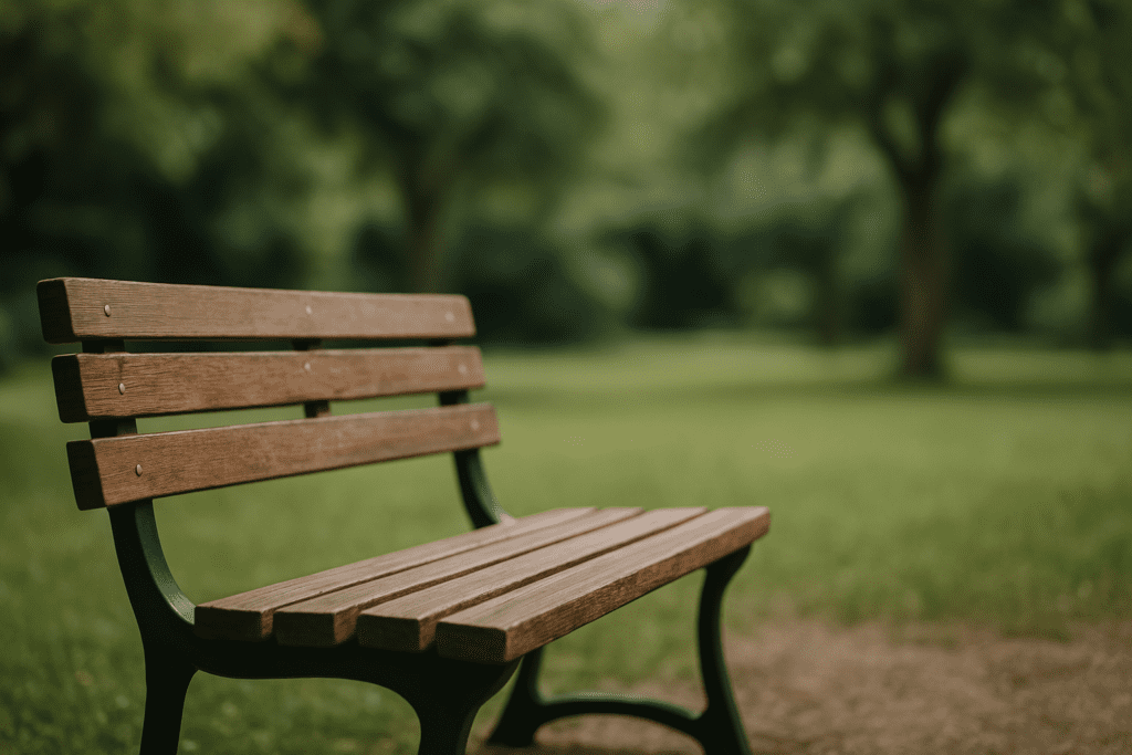 A wooden park bench with green metal supports sits empty on a gravel path, surrounded by a soft-focus grassy field and leafy trees under diffused natural light.